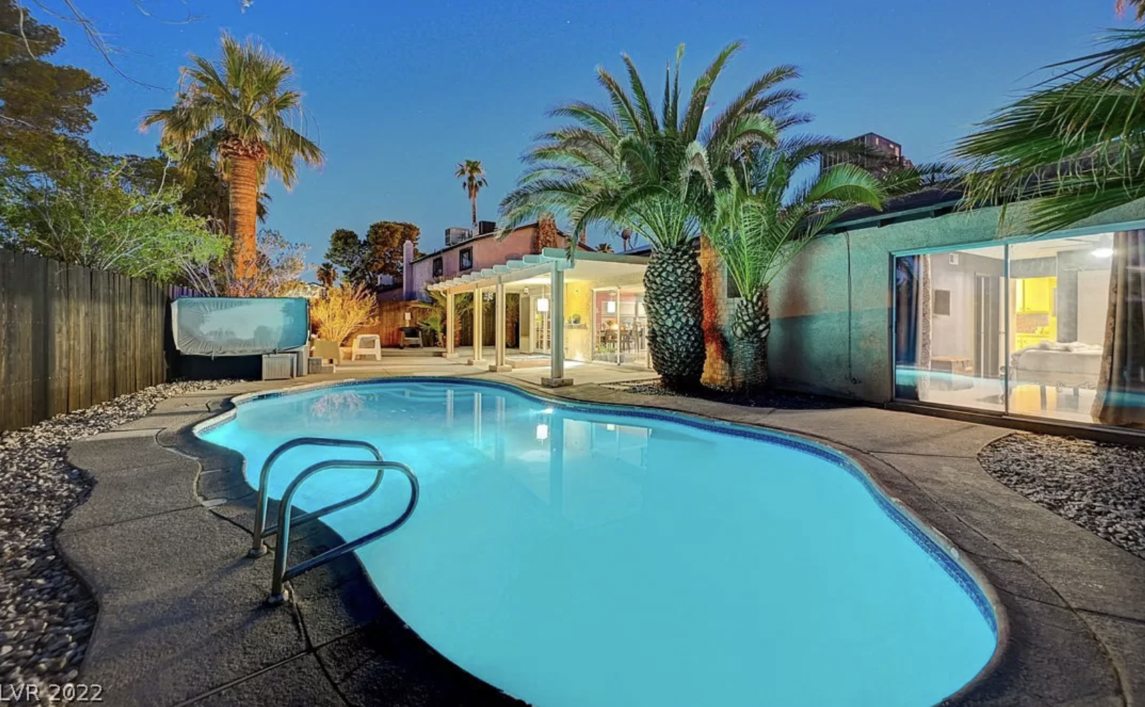 A colorful back yard with a bright blue pool, palm trees, covered patio, and large glass doors
